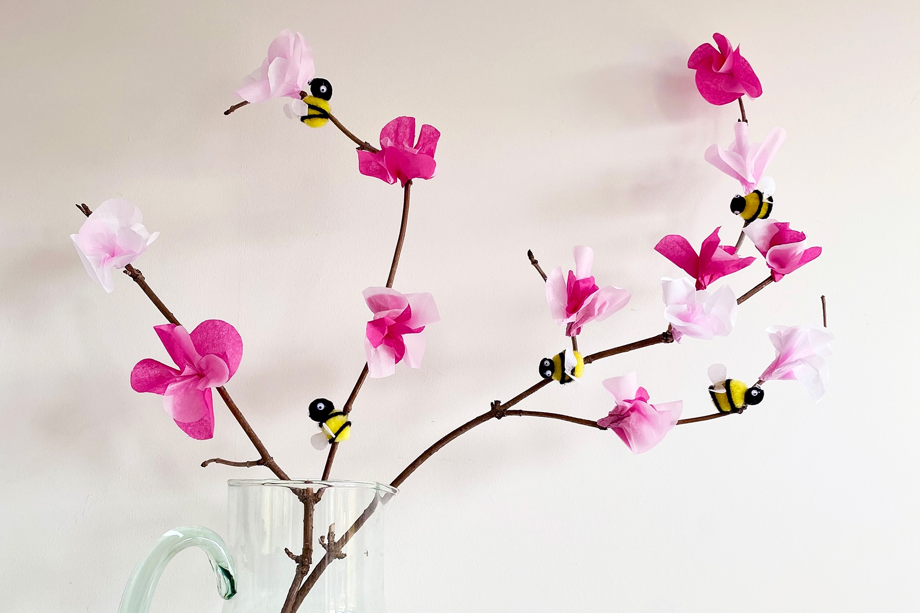 Photograph of tissue paper flowers and homemade pom pom bees stuck onto a tree branch and placed in a clear glass jug against a white background