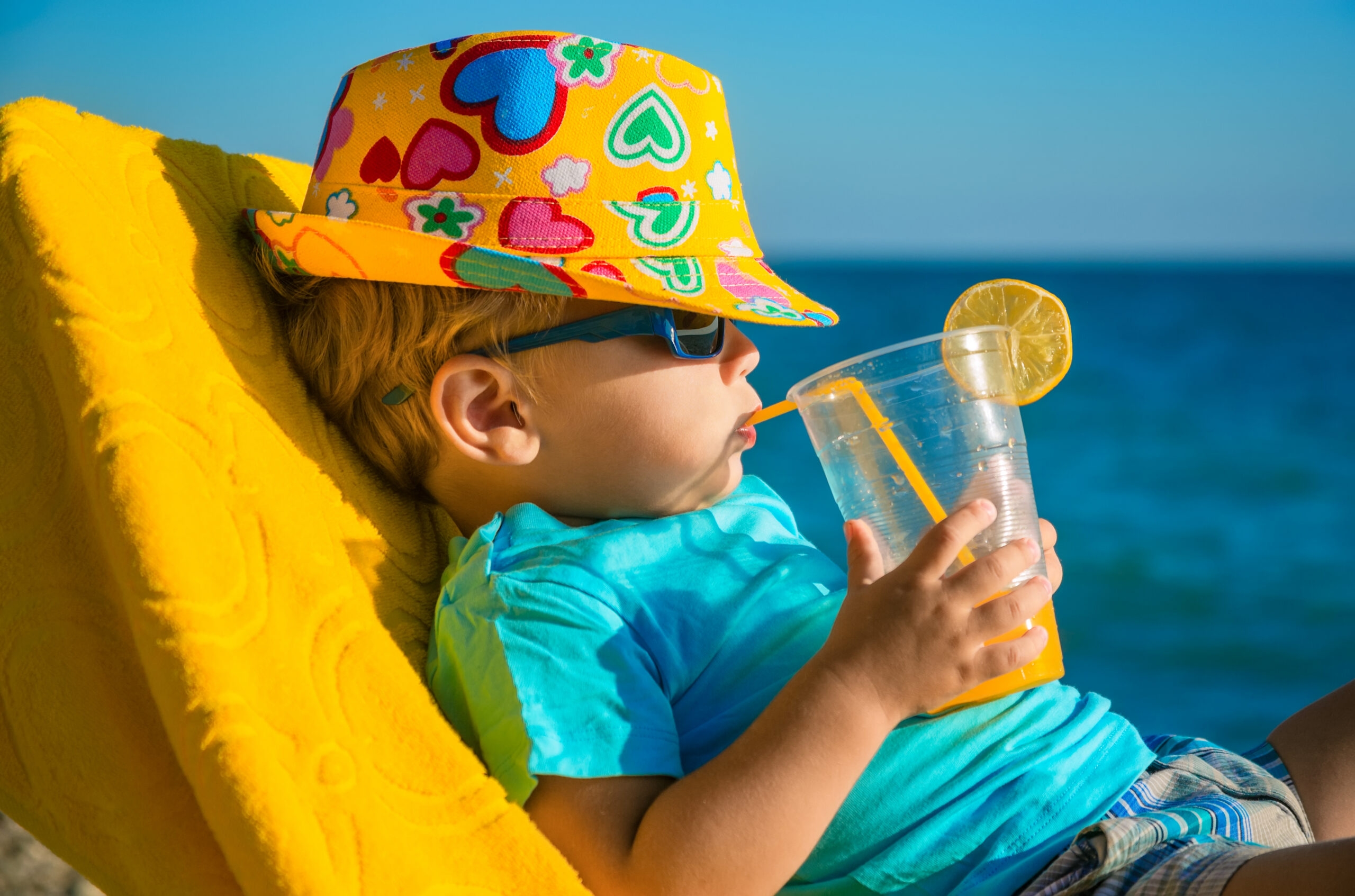 Boy kid in armchair with juice glass on beach against sea