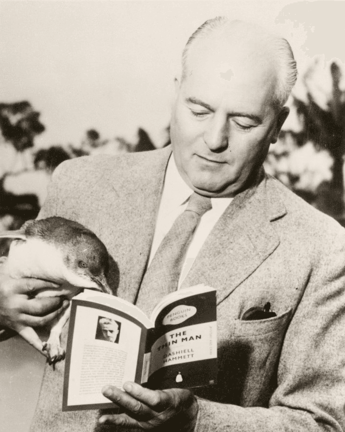 Photo of Sir Alan Lane holding a small penguin and reading a Penguin book