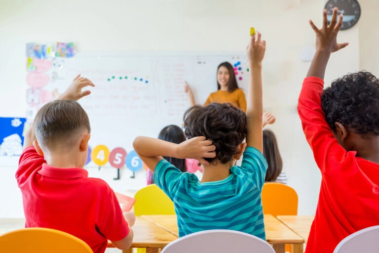 A photo of children in a classroom, raising their arms as the teacher has asked a question