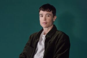 Photo of Elliot Page, Elliot sits in front of a green backdrop wearing a white t-shirt and green coat.