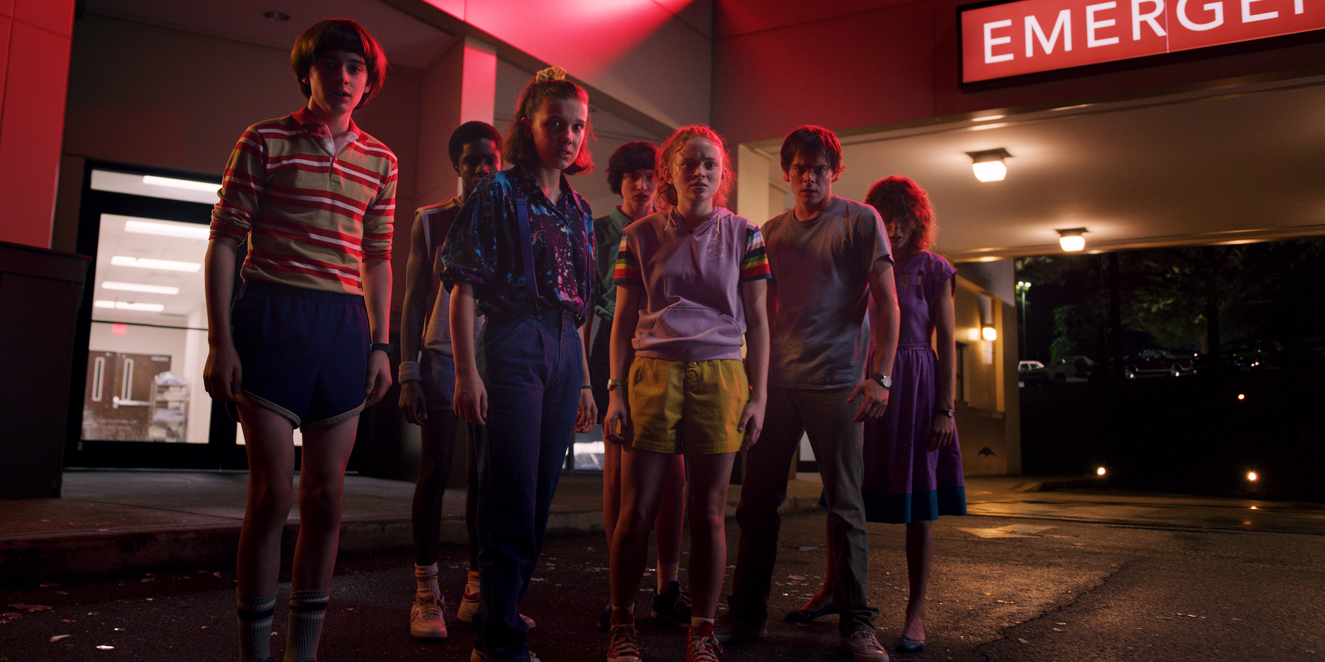 The teenage main characters from Stranger Things stand together in a group, in a shopping mall, lit by red neon lighting.
