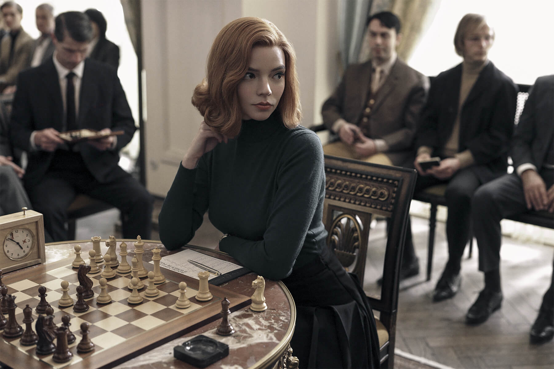 Anya Taylor-Joy as Beth Harmon in the Netflix series, The Queen's Gambit. Here Beth is seen looking pensive as a chess board, in the midst of a room of onlookers.