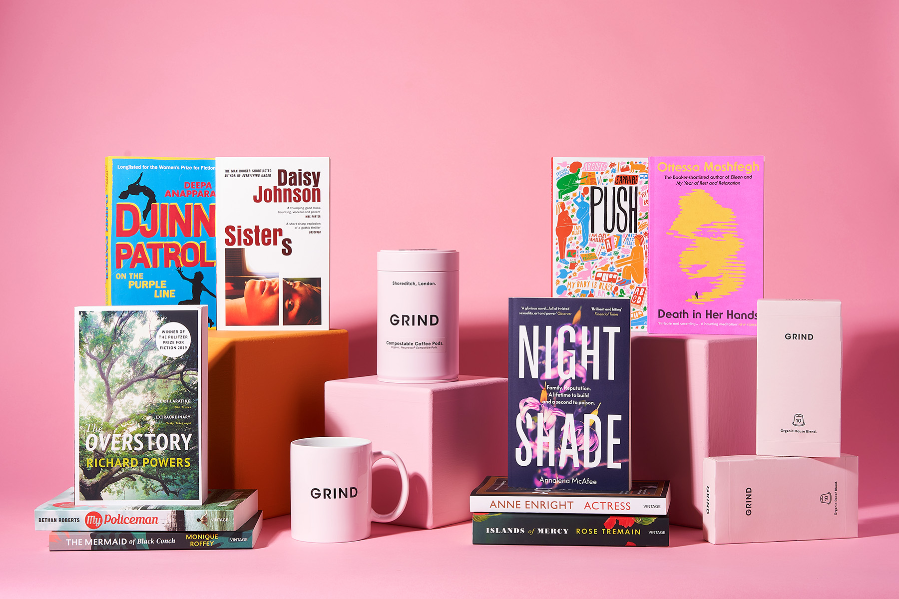 A photograph of 10 paperback books in stacks with Grind coffee tins, boxes and mugs against a pink background
