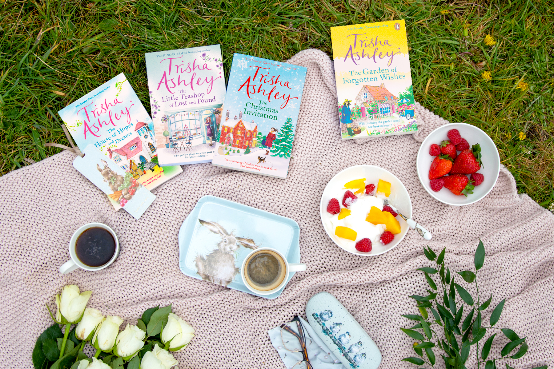 A photo of a variety of Trisha Ashley books on a picnic blanket with tea, fruit and more, on grass.