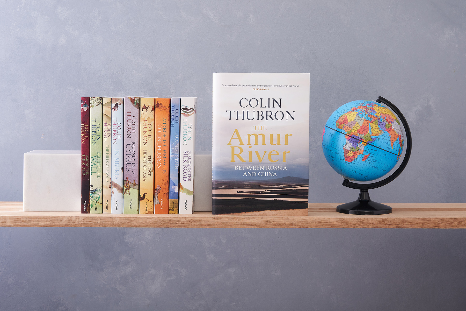 Colin Thubron's travel writing books standing upright on a shelf to show spines, with The Amur River turned to display its cover, alongside a small globe.