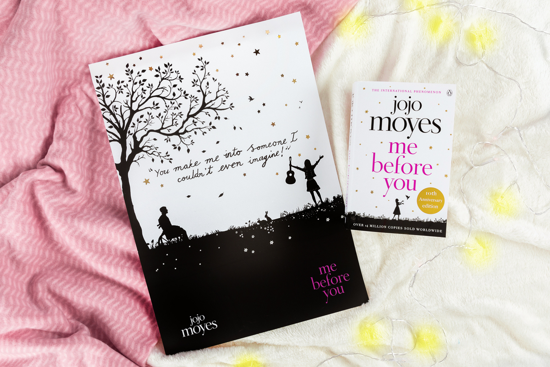 An image of Me Before You by Jojo Moyes on pink and white blankets, beside a string of yellow lights and a print from the book.