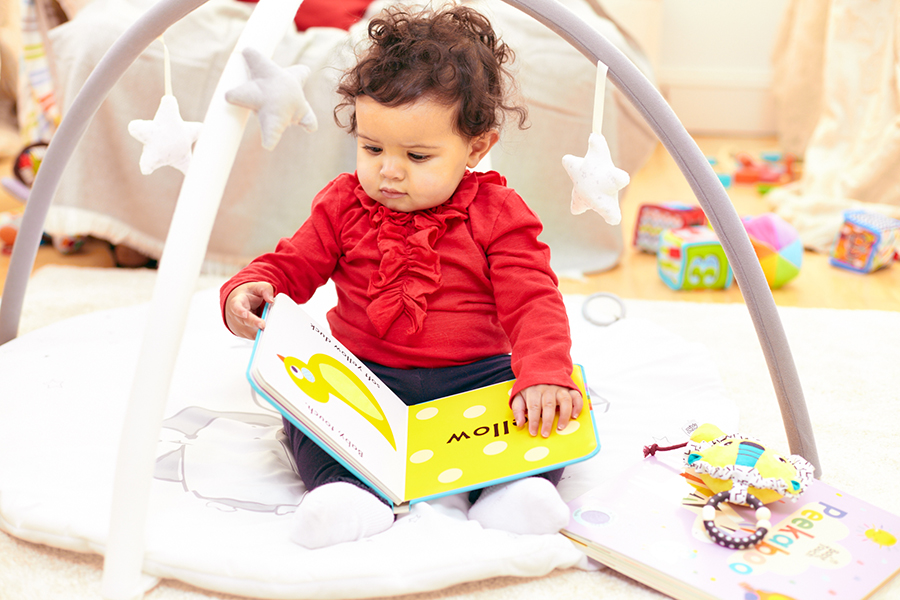 An image of a baby girl sitting on the floor with a board book in her lap. She is surrounded by other toys and books, and is trying to turn the page of the book she is holding