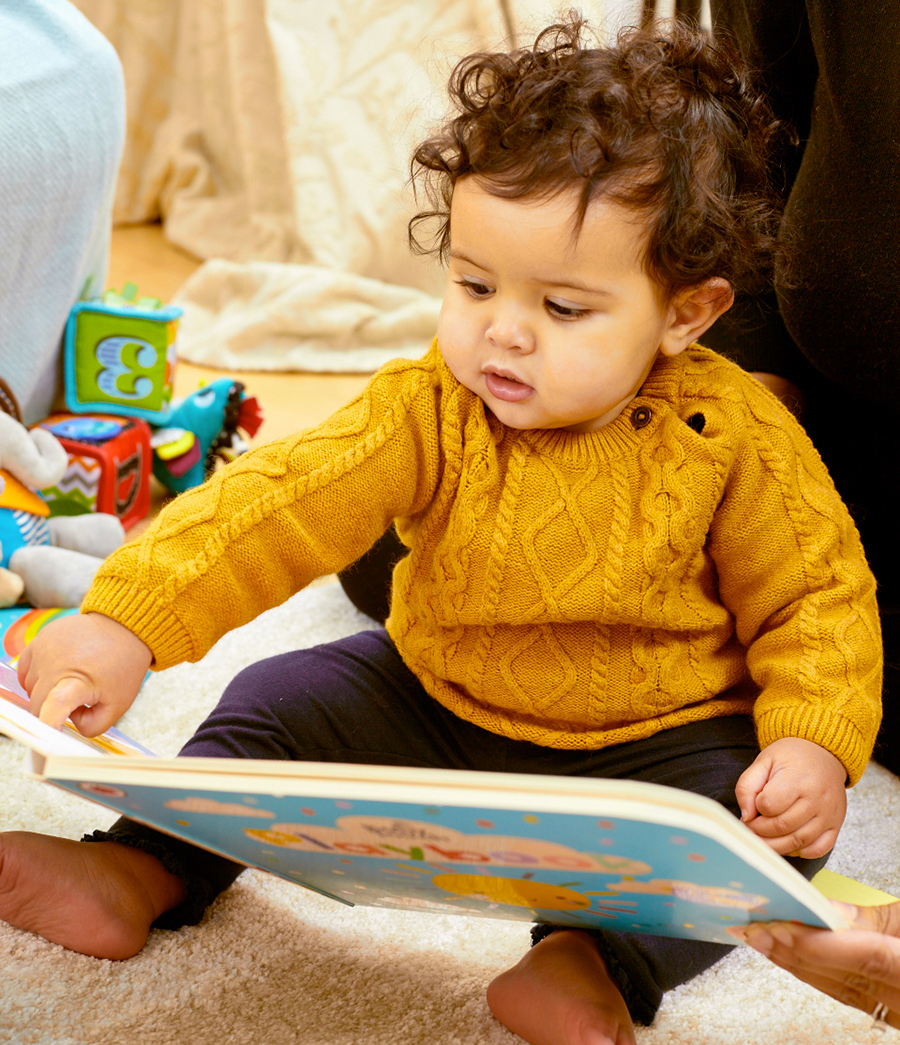 An image of a baby girl sitting on the floor with a board book being held in front of her by an adult. She is surrounded by other toys and books, and is pointing to something on the page of the book in front of her
