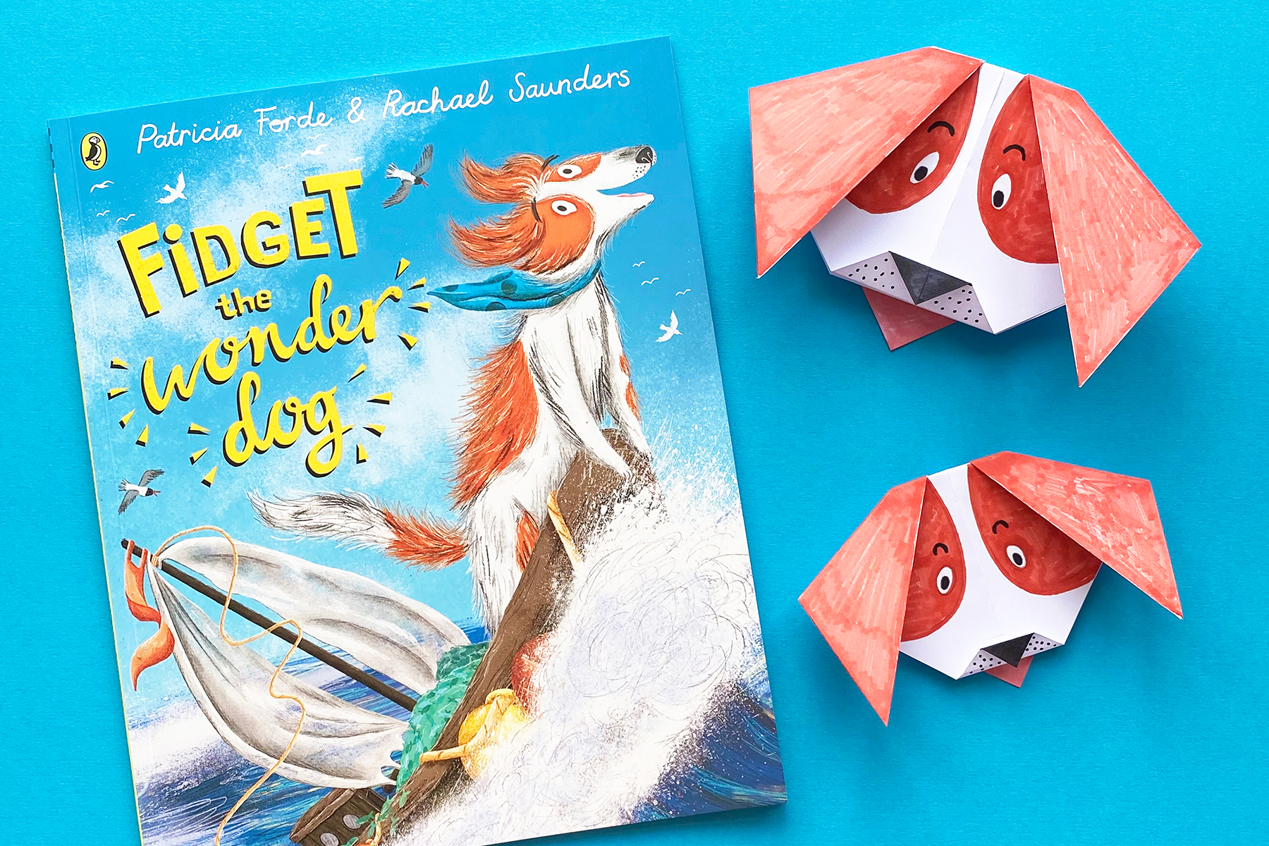 A photo of the book Fidget the Wonder Dog alongside a couple of origami Fidgets on a bright blue background