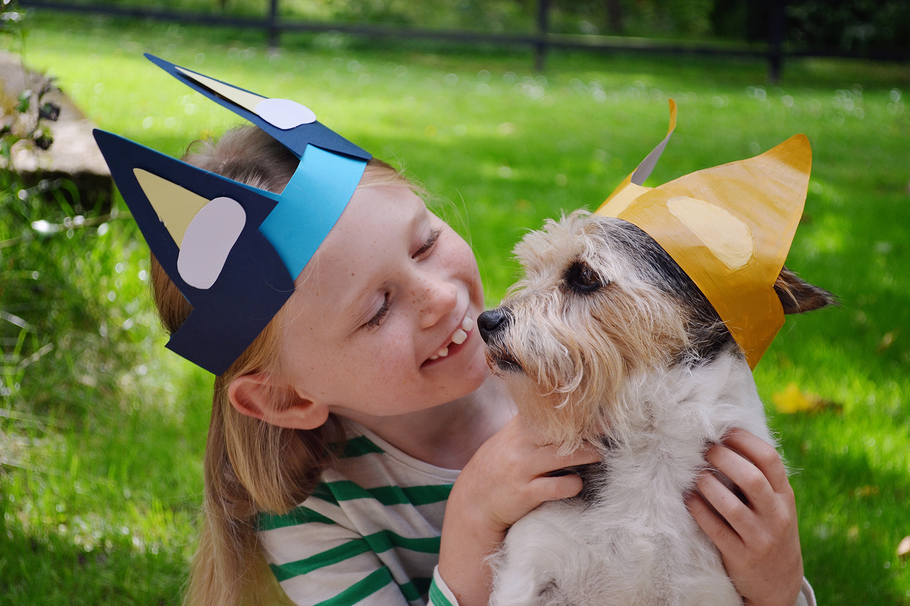 A photo of a girl sitting in a garden with a terrier type dog on her lap; they are both wearing a pair of Bluey and Bingo cut-out ears