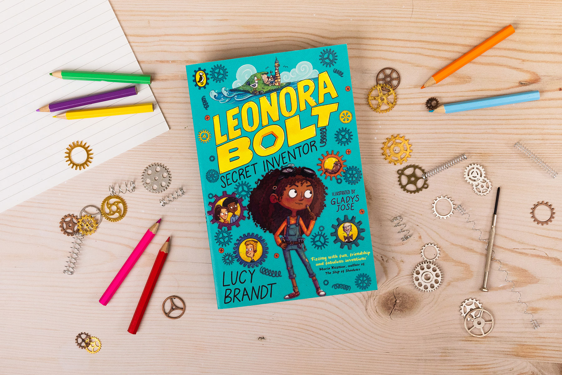 A photo of the book Leonora Bolt: Secret Inventor on a wooden table surrounded by colouring pencils, lined paper, screws and cogs