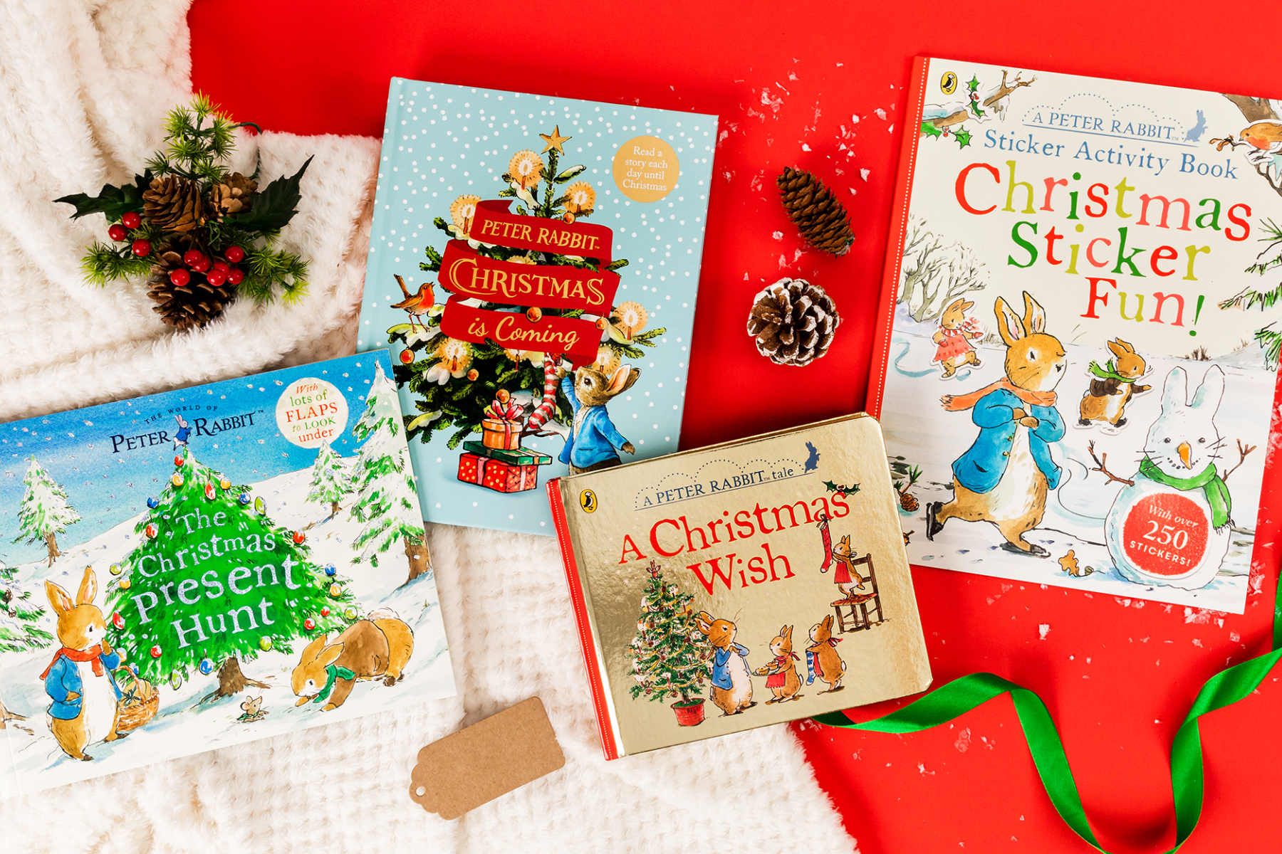 A photo of a bundle of Peter Rabbit books on a red background alongside a blanket, pine cones and green ribbon