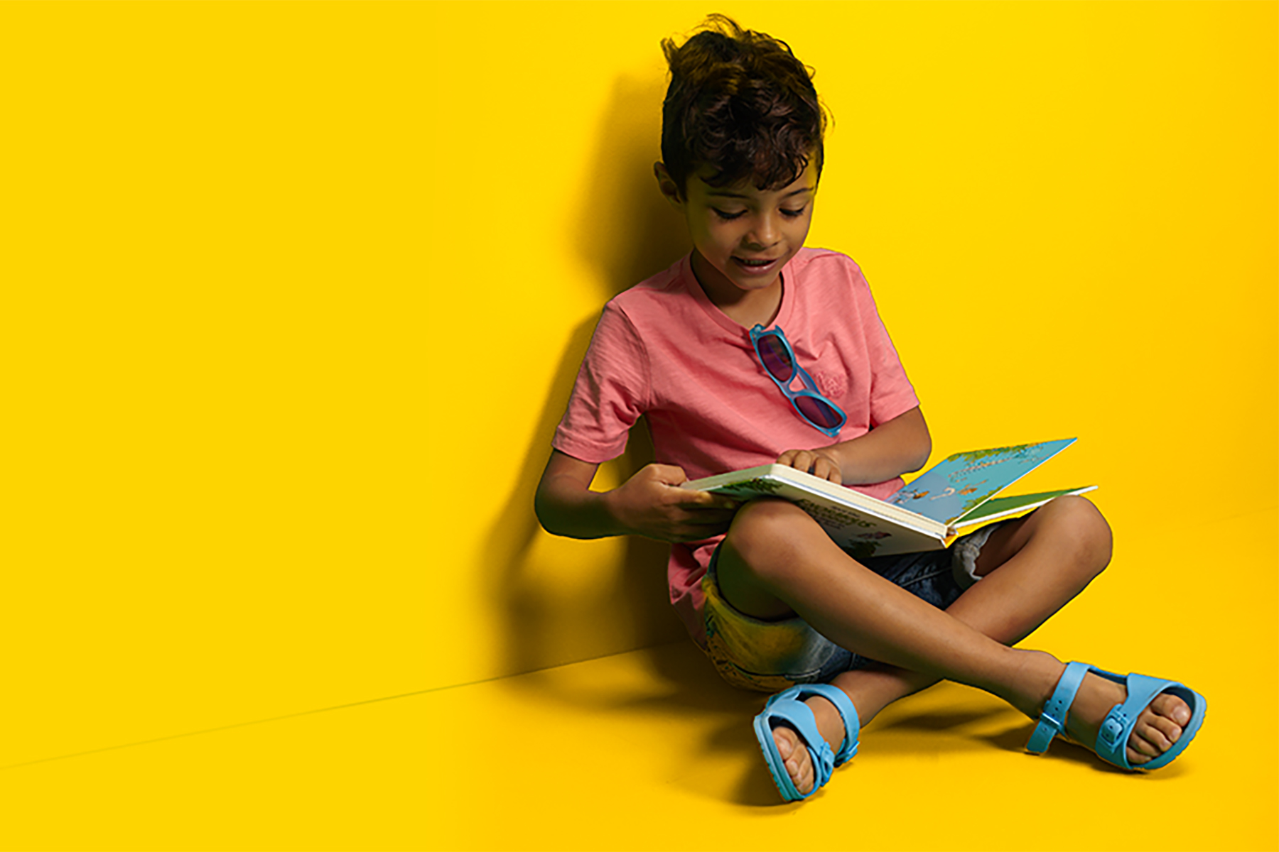 A photo of a young boy sitting against a bright yellow background whilst reading a book