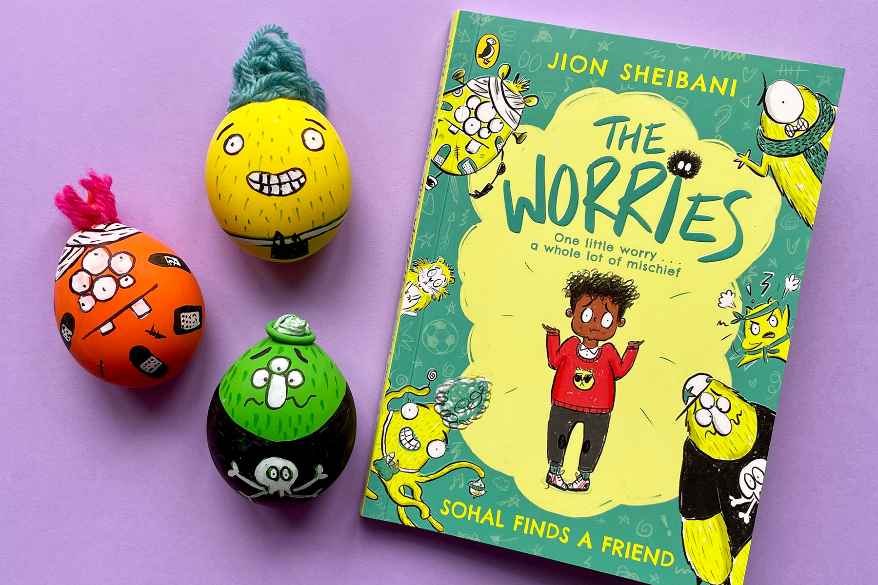 A photo of three stress balls with funny faces inspired by the characters in The Worries by Jion Sheibani; they are next to a copy of the book on a light purple background