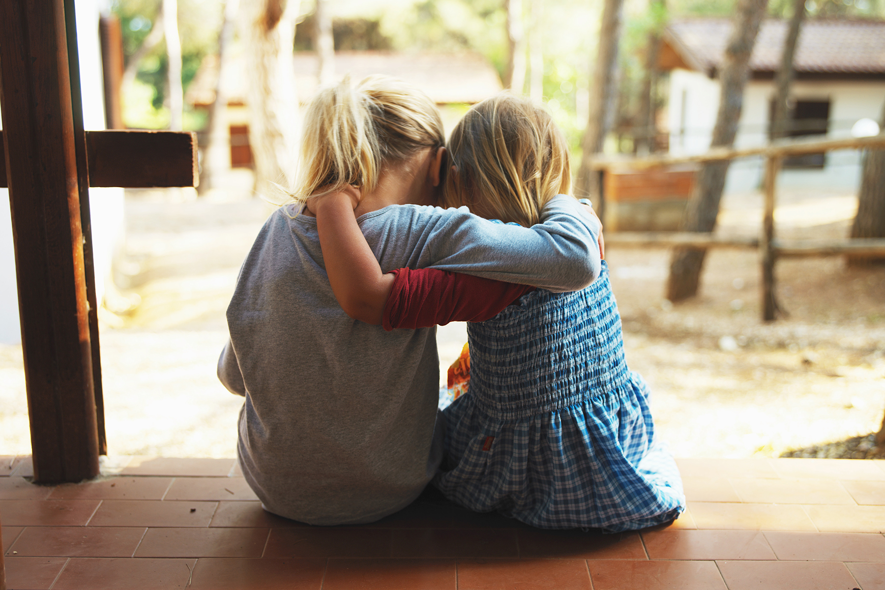 A photo of two young girls, sitting on a porch on a sunny day hugging and comforting each other.