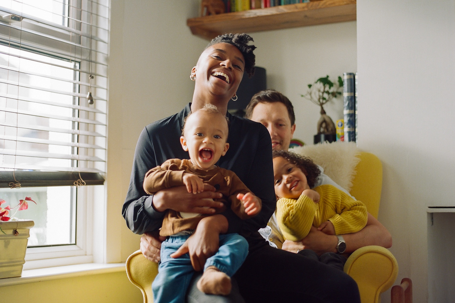 A photo of parents Ashleigh and Dan holding their two children; they're sitting on a yellow armchair in their home and all are laughing and smiling