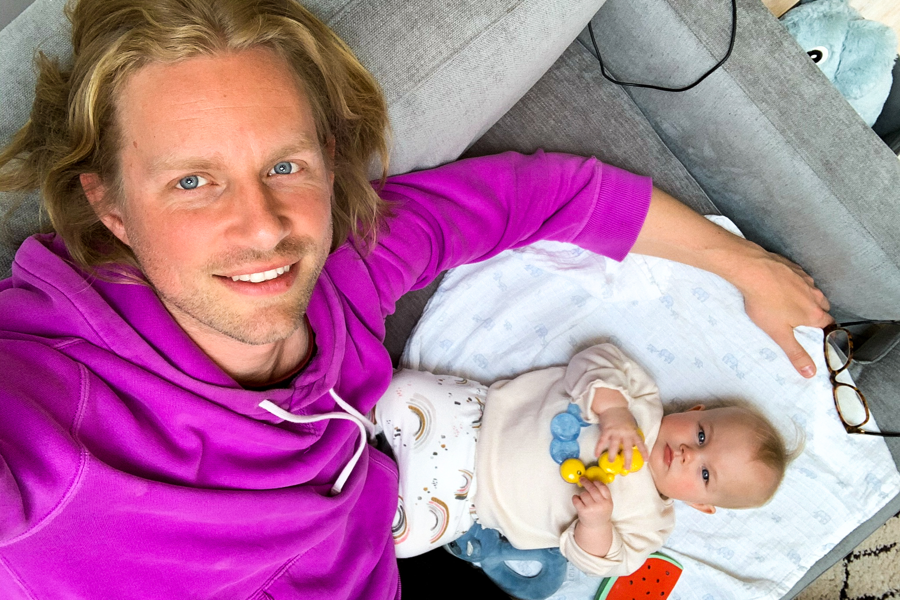 A photo of father Erik with his young baby girl; they are on a grey sofa; Erik is smiling and holding up the camera so the photo is a birds eye view