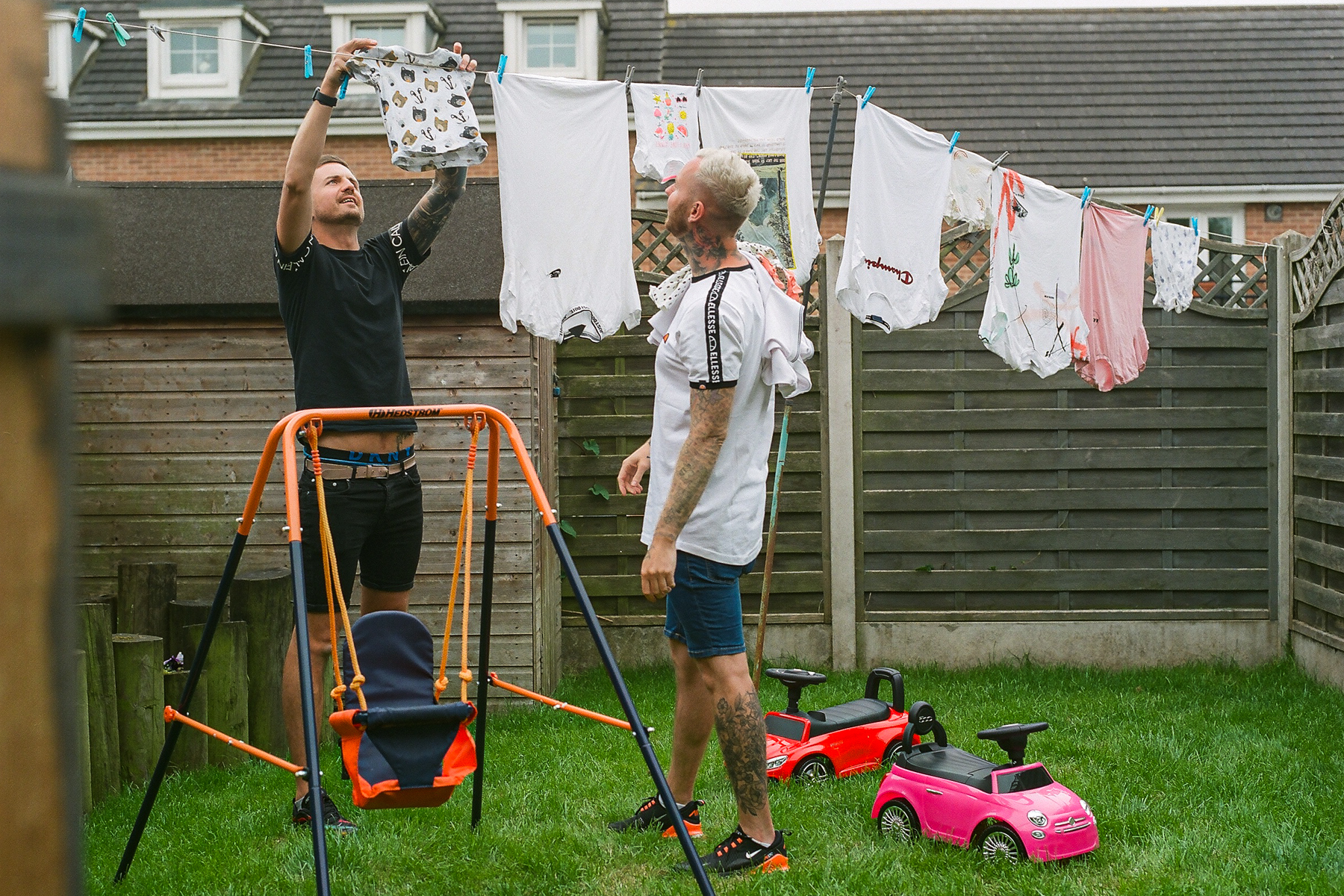 A photo of fathers Luke and Andy hanging clothes on a washing line in their back garden; they are surrounded by a swing and two toy cars