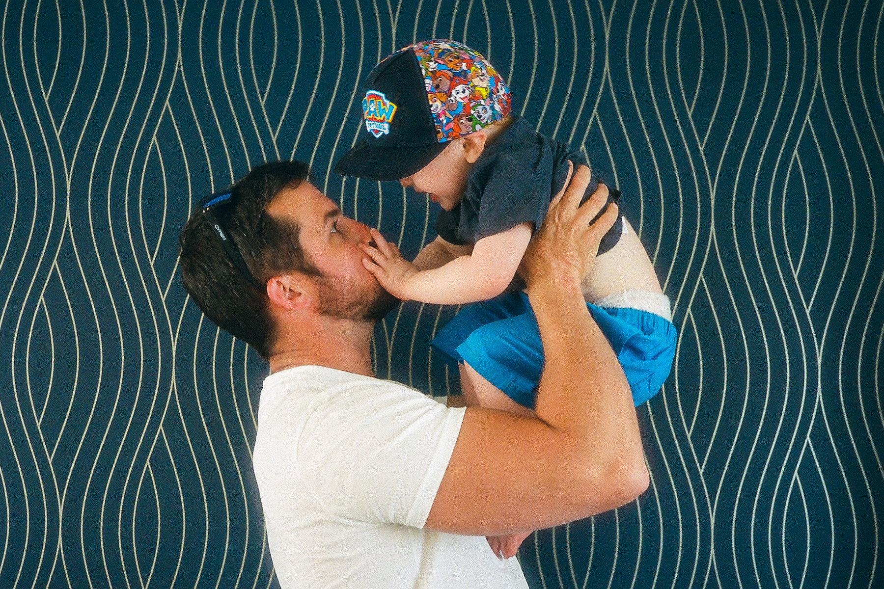 A photo of father Mark holding up his young son; they are against a navy and gold wallpaper backdrop; Mark's son's hands are on his mouth and Mark is making a silly face whilst his son giggles