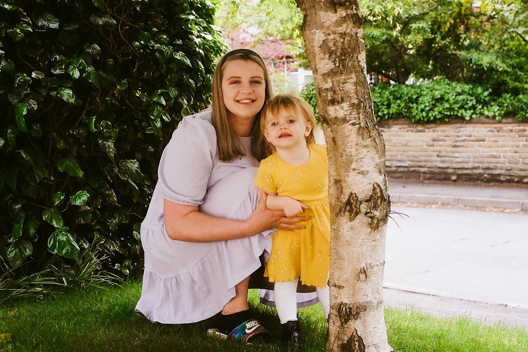 A photo of mum Megan smiling, crouched down by a tree with her daughter who is also looking into the camera; Megan is wearing a lilac dress and her daughter is wearing a yellow dress