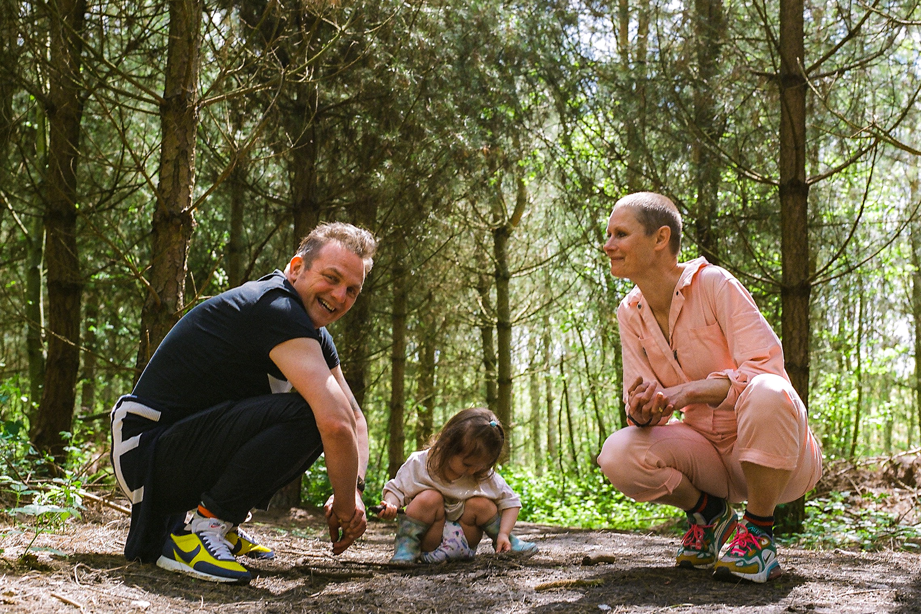 A photo of grandparents Nic and Mark with their grandchild; they are in a wooded area and all three at crouched down on the ground. Mark looks at the camera, Nic looks at Mark and their grandchild is playing with something on the ground