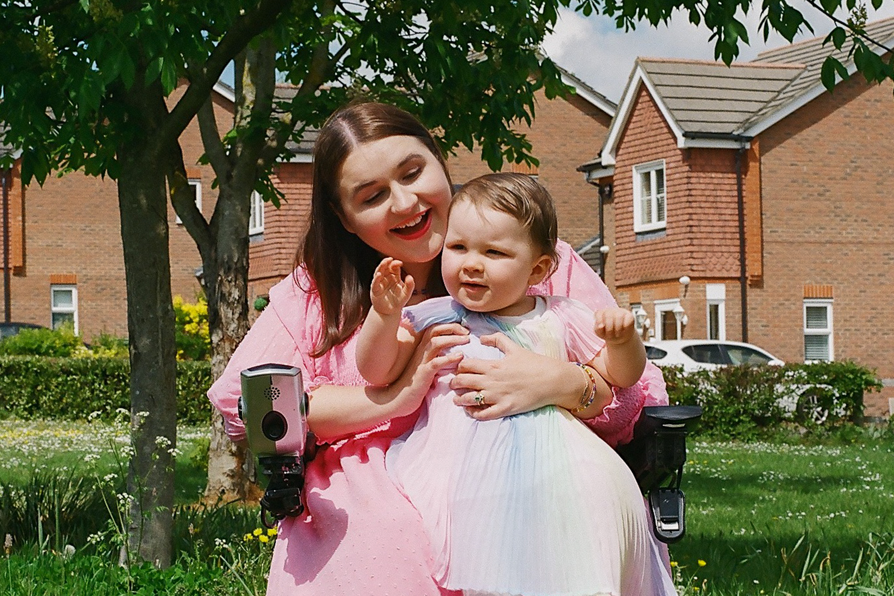 A photo of mother Sophie sitting in her wheelchair smiling and holding her young child against a backdrop of houses and greenery