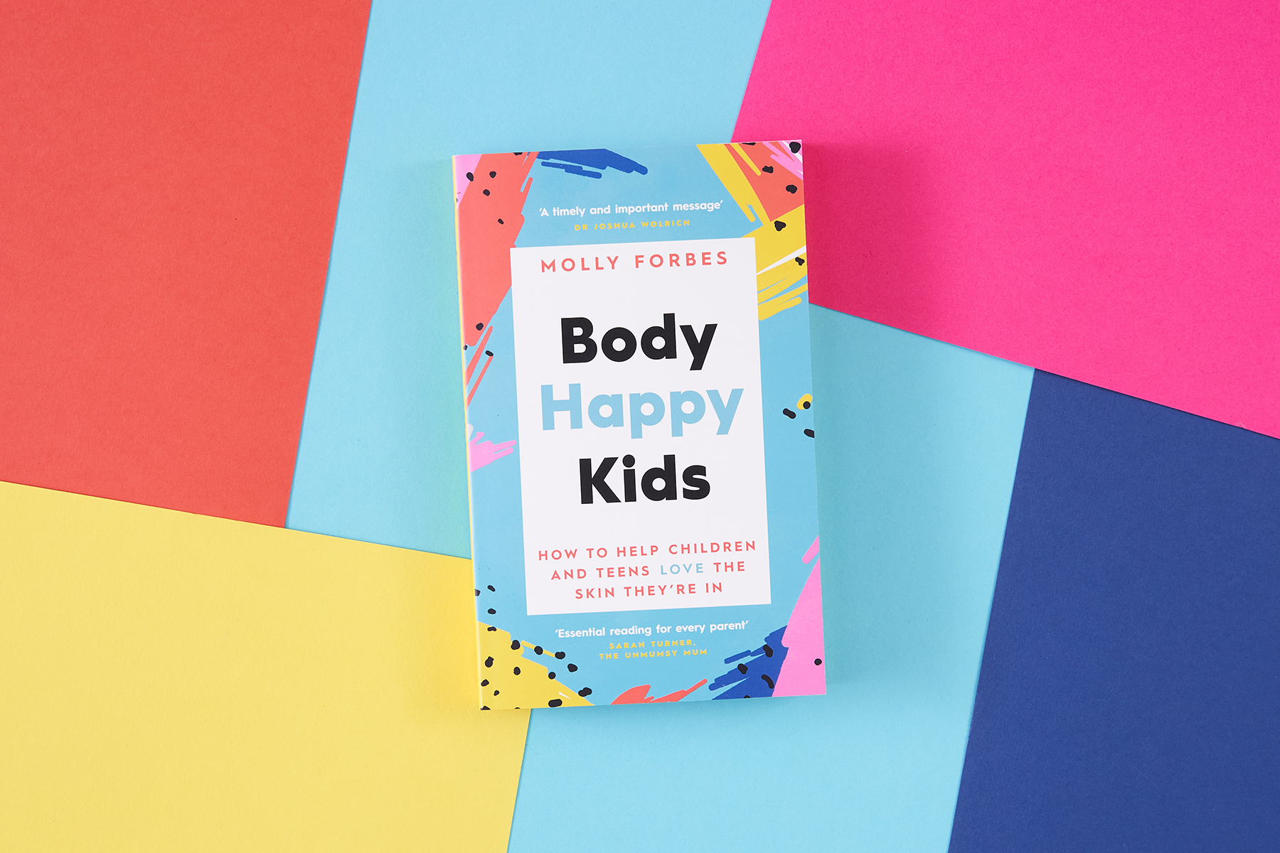 A photo of Molly Forbes' book Body Happy Kids on a multicoloured (coral, hot pink, dark blue, light blue and yellow) paper background
