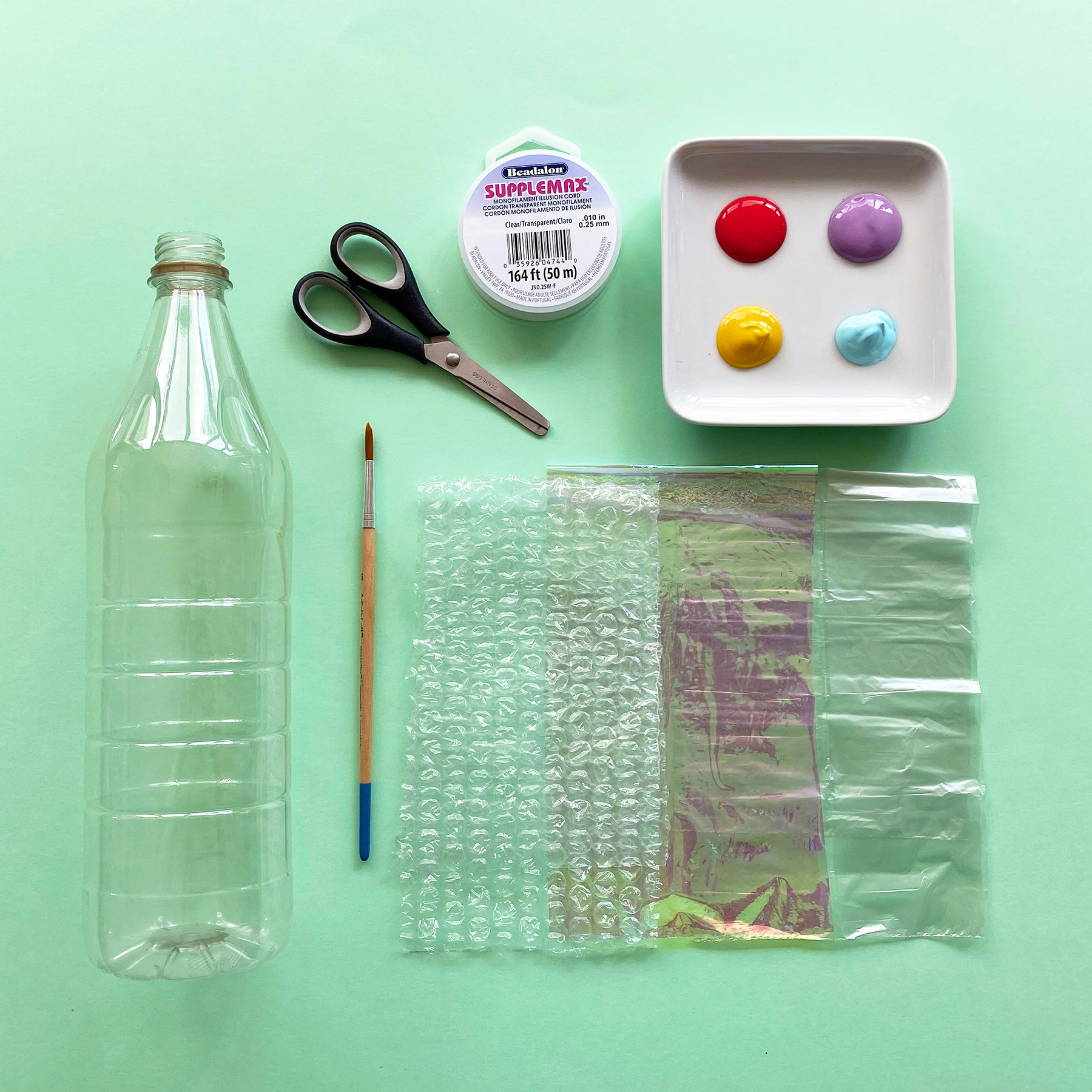A photo of all the things you will need to make a recycled bottle jellyfish including, a plastic bottle, small paintbrush, scissors, clear string, paints, cellophane and bubblewrap