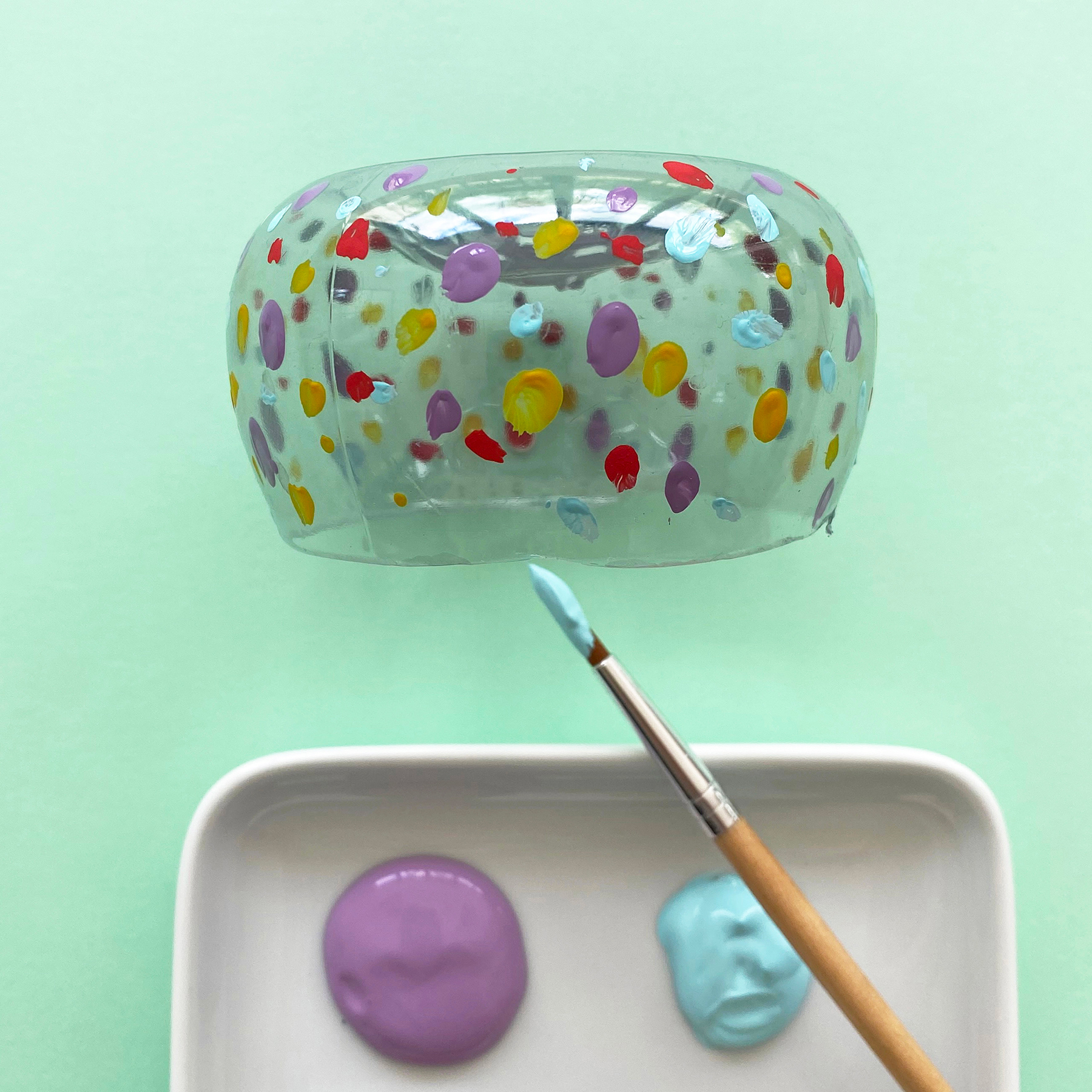 A photo of the bottom of a plastic bottle being painted in different colours alongside a dish of paints and a paintbrush on a mint green background