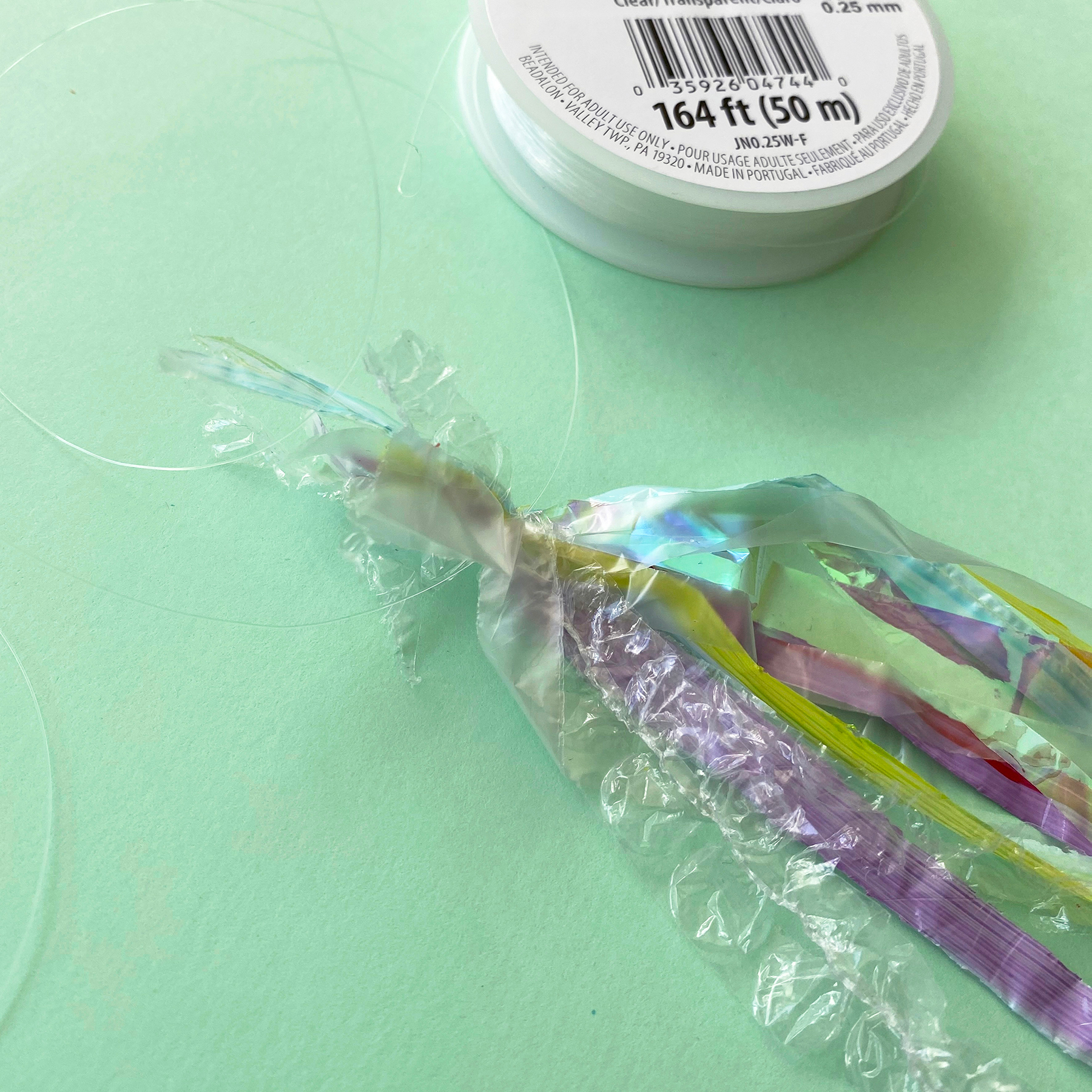 A photo of the plastic bag, cellophane and bubble wrap strips being tied together using clear string on a mint green background