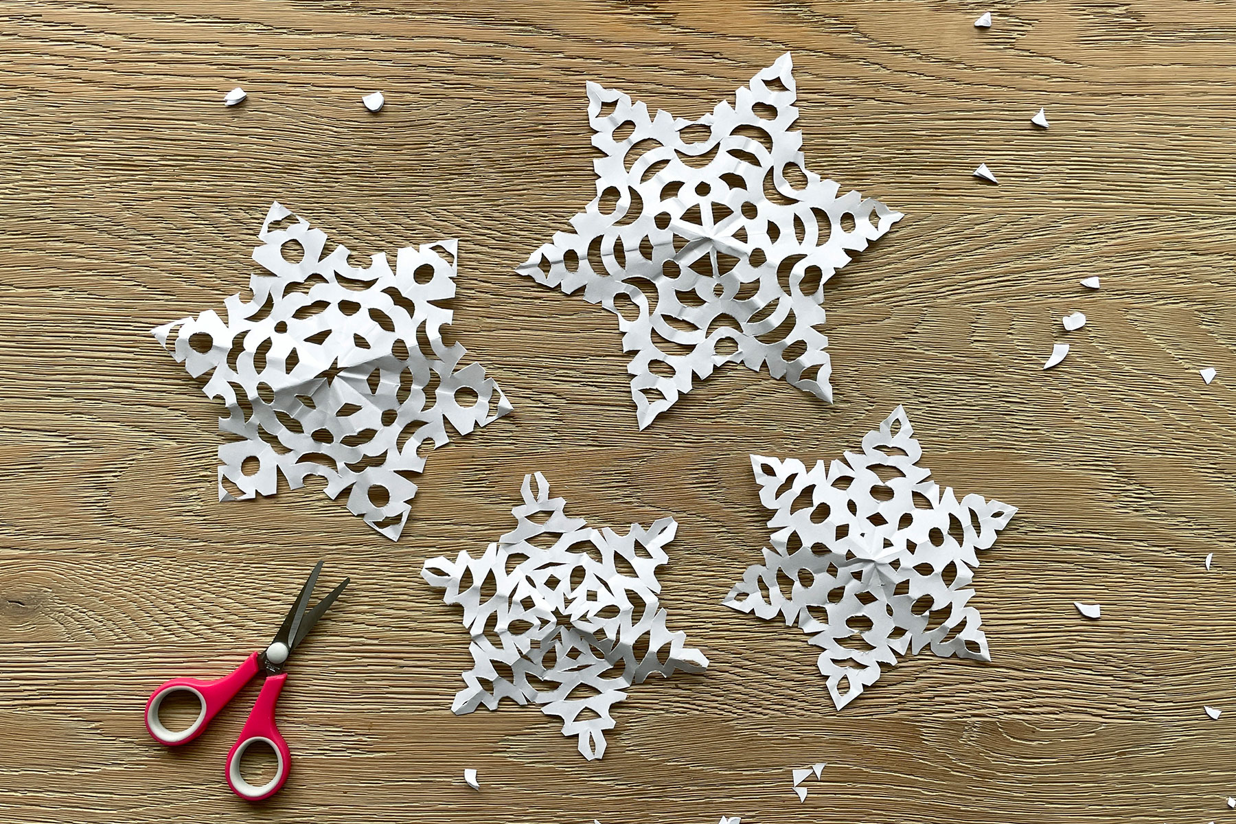 A photo of four paper snowflakes laid out next to each other on a wooden table alongside a pair of scissors