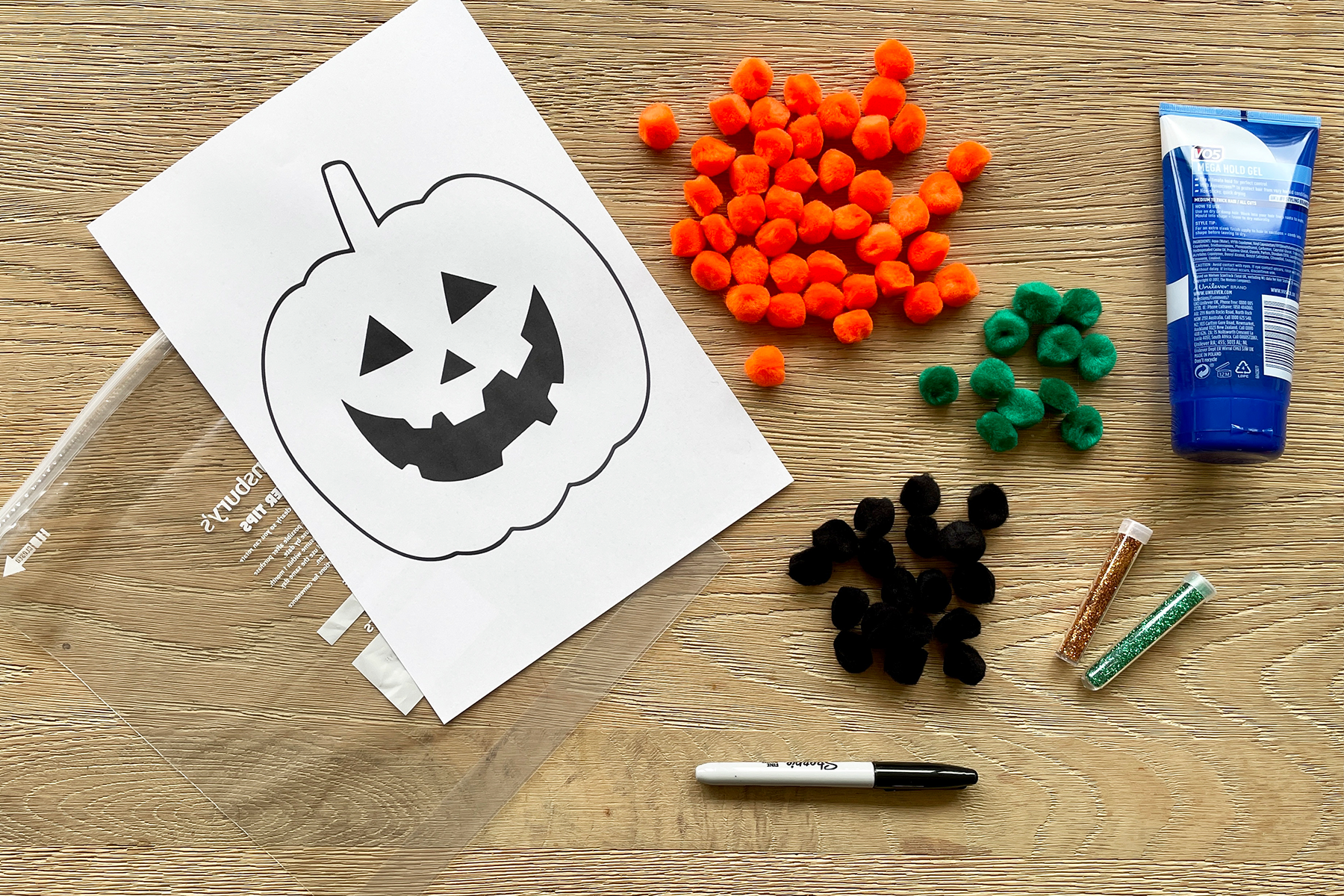 A photo of a pumpkin stencil on top of a plastic bag with orange, green and black furry pom poms, glitter and clean hair gel